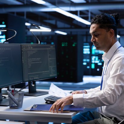 Person arbeitet an einem Schreibtisch mit zwei Monitoren in einem modernen Büro mit Servern im Hintergrund.