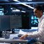 Person arbeitet an einem Schreibtisch mit zwei Monitoren in einem modernen Büro mit Servern im Hintergrund.