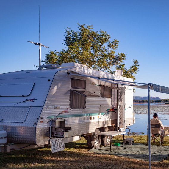 TÜV NORD Gasprüfung Zwei Personen sitzen an einem See bei blauem Himmel. Ein Wohnwagen steht auch auf der Wiese.