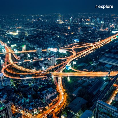 Stadtansicht bei Nacht Eine lebendige Stadtlandschaft bei Nacht, die komplexe Autobahnkreuzungen und beleuchtete Gebäude zeigt. Die Straßen sind von Lichtern der Fahrzeuge erhellt, und der Hashtag "#explore" ist oben rechts im Bild zu sehen.