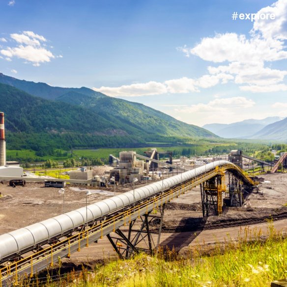 Industrieanlage mit Rohrleitung in bewaldetem Bergtal Industrieanlage in einem breiten Tal mit bewaldeten Bergen im Hintergrund. Eine lange, über Gestellen geführte Rohrleitung oder Förderstrecke verläuft diagonal durch das Gelände zu mehreren Industriegebäuden und technischen Anlagen. Der Himmel ist blau mit vereinzelten Wolken.