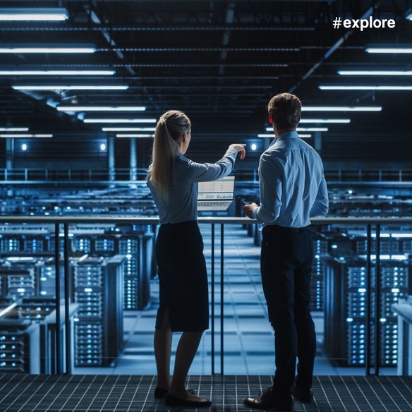 Analyse von Daten im Rechenzentrum. Zwei Personen stehen in einem modernen Rechenzentrum und betrachten einen Laptop mit Datenanalysen. Die Umgebung ist von blauen Lichtern und zahlreichen Servern geprägt, die eine technologische und futuristische Atmosphäre schaffen.