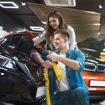 Elektromobilität Ein Mann kniet und eine Frau steht neben ihm in einer Halle mit Autos. Die Haube vorne ist bei einem Auto offen und der Mann hält einen gelben Schlauch in der Hand