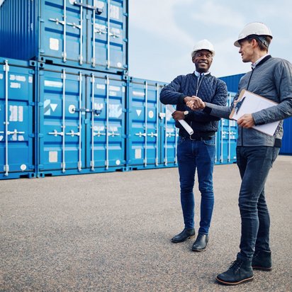 Zwei Personen an einem Umschlagplatz für Handelswaren Symbolbild IFS Broker Zertifiizierung - Zwei Männer mit weißen Helmen und legerer Kleidung mit Klemmbret vor Kisten mit Ware