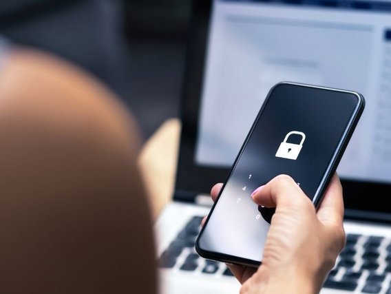 Ein Smartphone mit weißem Schloss-Symbol und Zifferntastatur auf dem Bildschirm, gehalten von einer Person vor einem Laptop, das Mobilgeräte-Sicherheit darstellt.