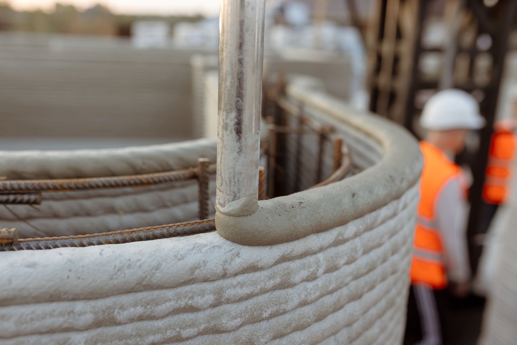 Detailaufnahme einer geschwungenen 3D-gedruckten Betonwand mit Bewehrungsstahl und einem Bauarbeiter im Hintergrund.