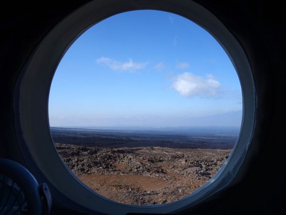 Blick durch ein rundes Fenster auf eine karge, rötlich-braune Vulkanlandschaft unter blauem Himmel mit weißen Wolken.