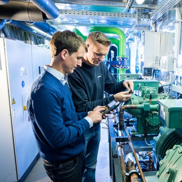 Inspektion im Maschinenraum eines Schiffes Zwei Mitarbeitende stehen im Prüfbereich der Kälte-, Klima- und Lüftungstechnik bei TÜV NORD und blicken auf technische Geräte, die sie in den Händen halten.