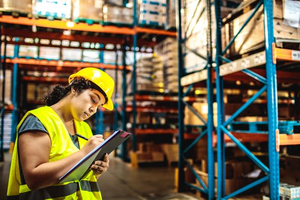 Lagerlogistik und Bestandsmanagement im Überblick Mitarbeiterin mit Schutzweste und Helm dokumentiert Lagerbestände in einem großen Hochregallager