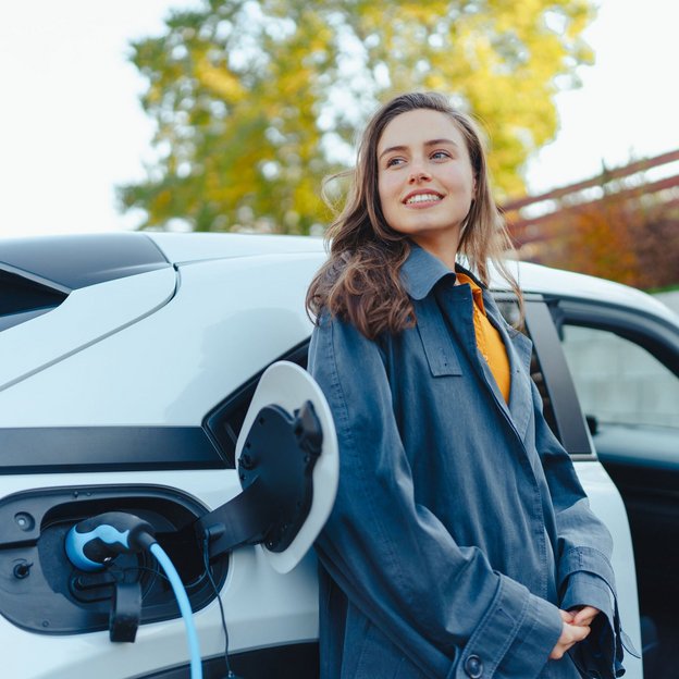 Elektromobilität als Schritt in eine nachhaltige Zukunft. Frau steht neben einem weißen Elektroauto, das an einer Ladesäule angeschlossen ist.