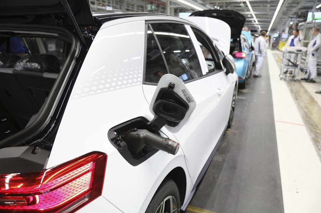 Weißes Elektroauto in einer Fertigungshalle mit geöffneter Ladeklappe und Ladestecker, Fabrikarbeiter im Hintergrund.