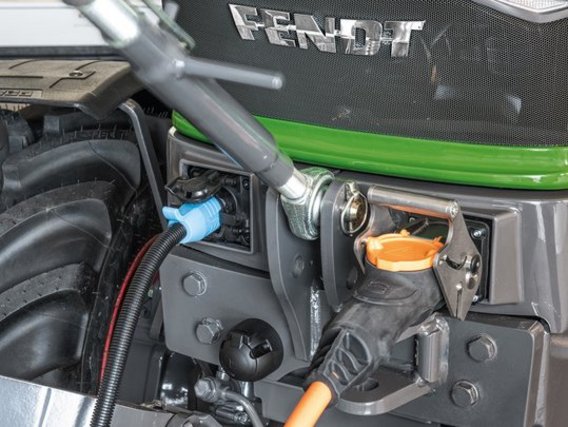 Nahaufnahme des Motorraums eines Fendt-Traktors mit grüner Lackierung, zeigt verschiedene Anschlüsse und Wartungsöffnungen. Sichtbar sind blaue und orange Verschlüsse sowie technische Komponenten der landwirtschaftlichen Maschine.