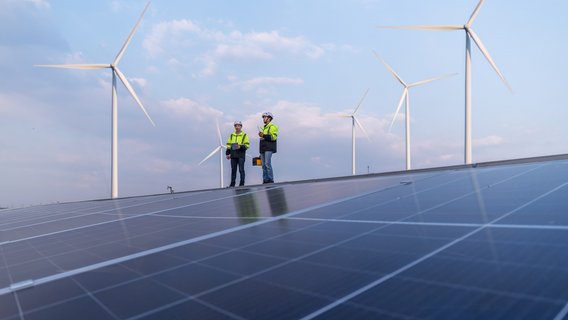 Inspektion von Photovoltaik- und Windenergieanlagen durch TÜV-Fachkräfte Zwei TÜV NORD-Ingenieure prüfen Solarmodule vor Windrädern unter blauem Himmel
