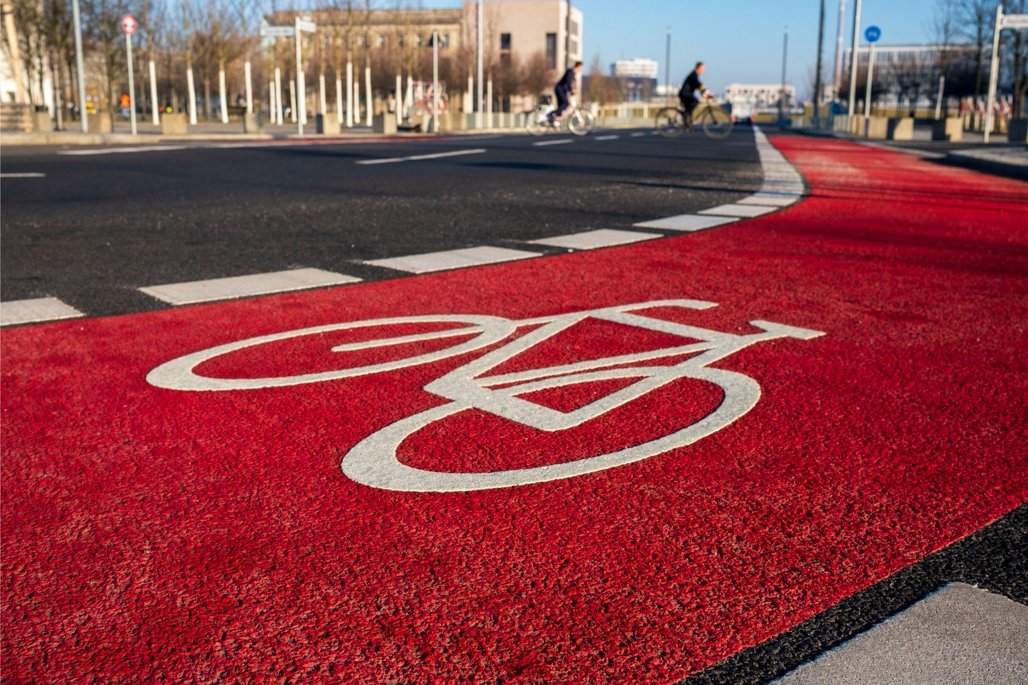 Fahrradweg an einer Kreuzung Ein roter Fahrradweg mit einem weißen Fahrradsymbol führt über eine Kreuzung. Im Hintergrund sind Radfahrer und städtische Gebäude zu sehen. Die Szene ist bei Tageslicht aufgenommen.