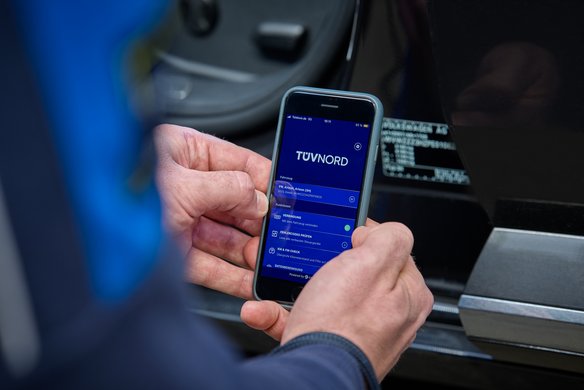 Eine Hand hält ein Smartphone mit der TÜV Nord App, die Fahrzeugdaten eines VW Arteon anzeigt, vor einem schwarzen Auto.