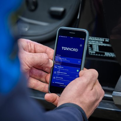 Eine Hand hält ein Smartphone mit der TÜV Nord App, die Fahrzeugdaten eines VW Arteon anzeigt, vor einem schwarzen Auto.