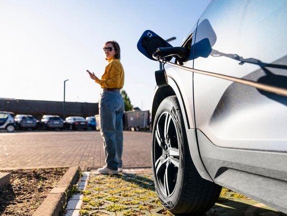 TÜV NORD-Ladesäule Frau steht neben einem Elektroauto an einer Ladestation und nutzt ihr Smartphone.